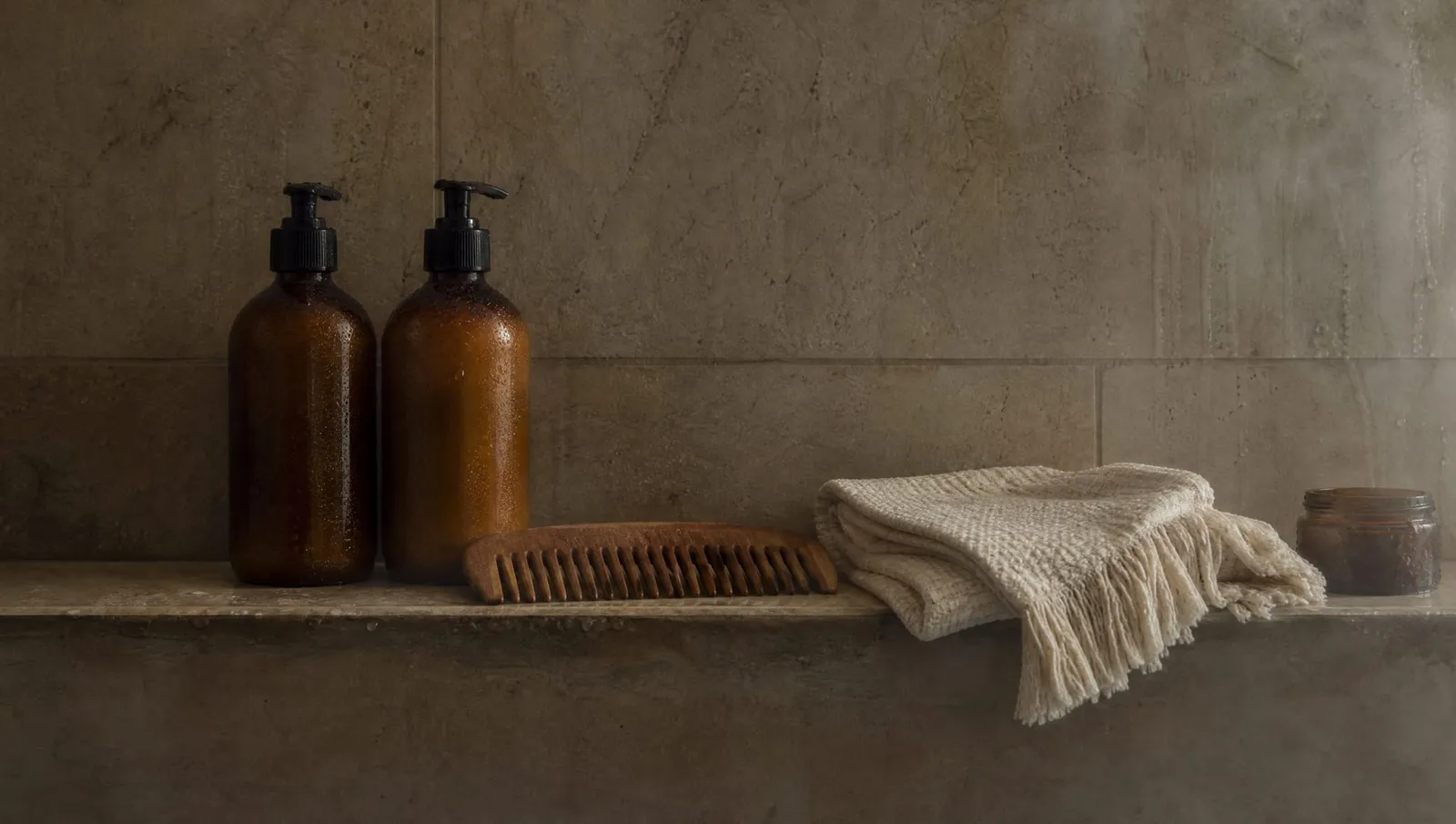 Two amber pump bottles, a wooden comb, a folded cream cotton towel and a small jar on a tiled ledge, with steam on the wall behind