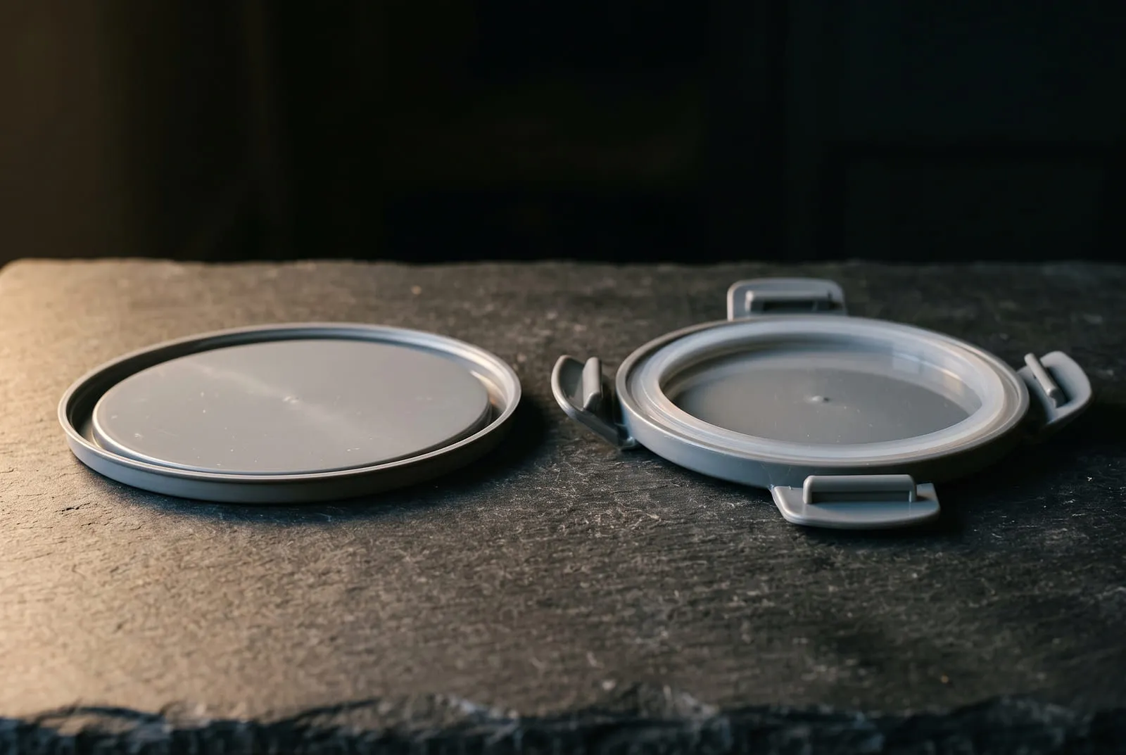 Two container lids photographed side by side on dark slate: on the left a plain thin press-on plastic lid lying flat with no gasket, on the right a rigid lid with a soft silicone gasket ring seated in a channel and four hinged locking tabs folded down over the rim