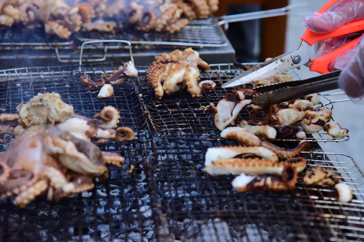 Close-up of grilled octopus being prepared with scissors on outdoor barbecue.